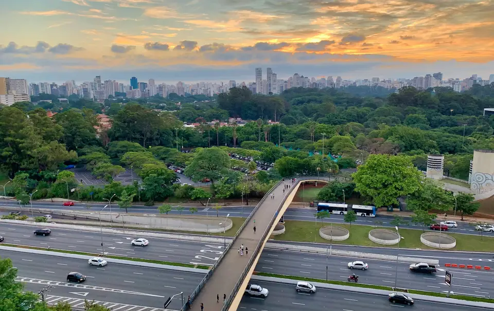 Vista do alto de avenidas movimentadas de São Paulo ao entardecer, com viaduto, árvores e prédios ao fundo