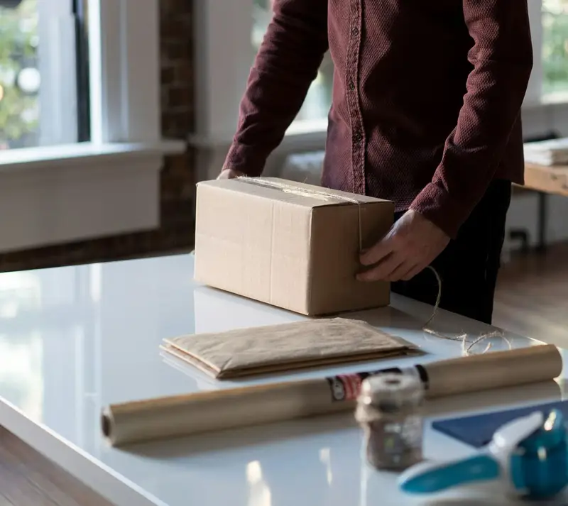 Pessoa fechando uma caixa de papelão sobre a mesa, com papel pardo e barbante ao lado