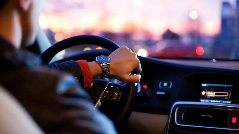 Mãos de um motorista no volante, com o painel do carro e a rua desfocada ao entardecer