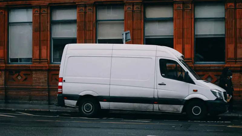 Van branca de carga estacionada em uma rua de cidade, em frente a um prédio de tijolos